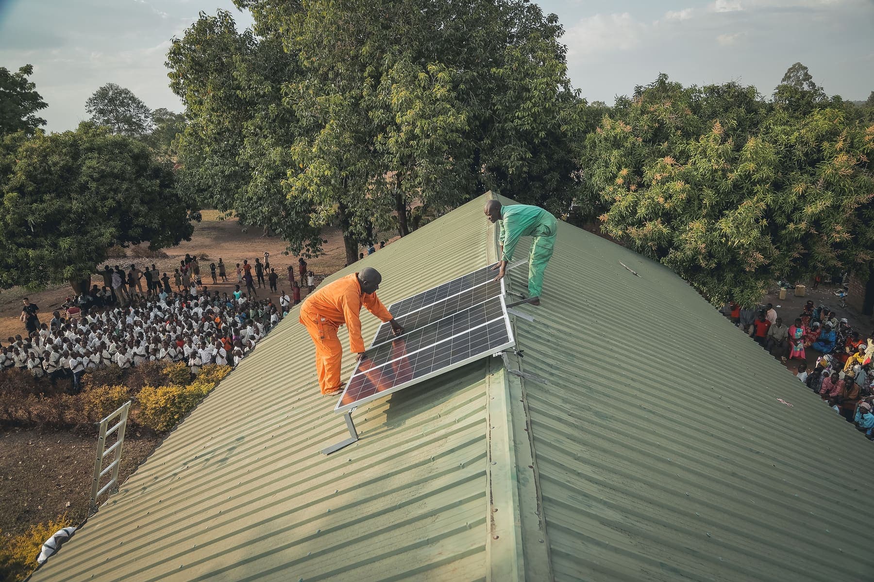 Two technicians fitting a solar panel on a green iron-sheet roof while a crowd watches below